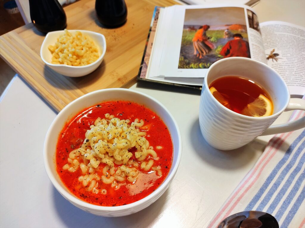 Stół kuchenny, a na stole miseczka zupy pomidorowej, kubek herbaty, książka i akcesoria kuchenne. Stół gotowy do kolacji. 