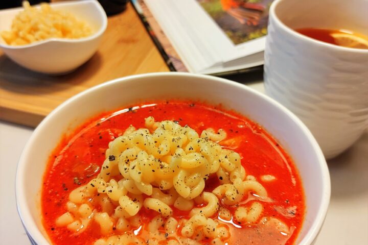Na stole miseczka zupy pomidorowej, kubek z herbatą, akcesoria kuchenne i książka.