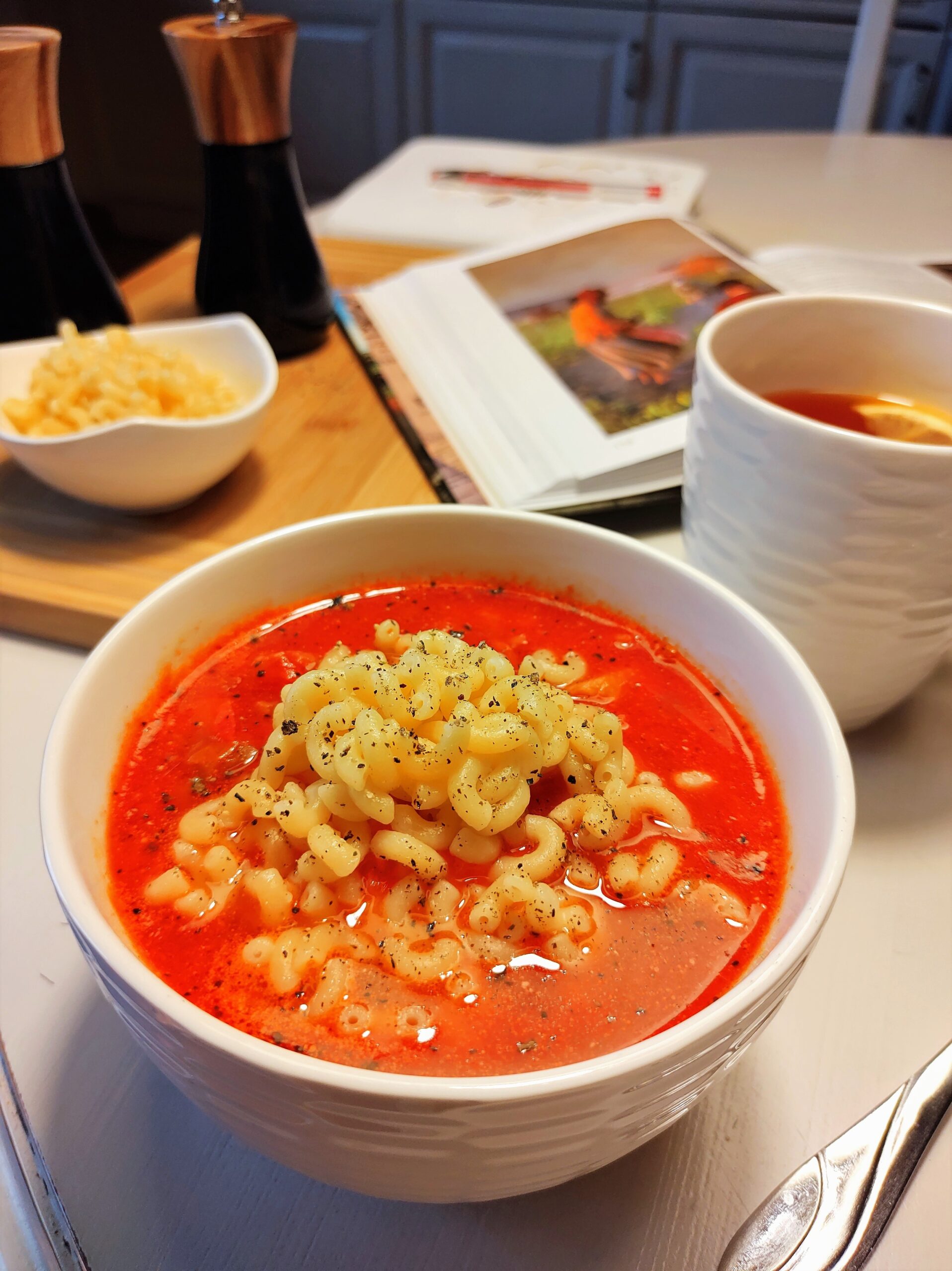 Na stole miseczka zupy pomidorowej, kubek z herbatą, akcesoria kuchenne i książka.